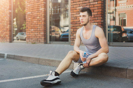Young Man Resting on Sidewalk With Water Bottle in Urban Setting During Daylight