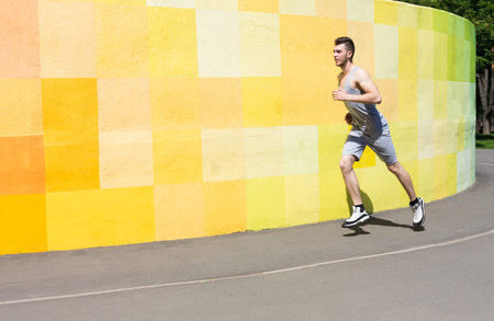 Young Man Runs on Path Beside Bright Yellow and Orange Wall on Sunny Day in Park