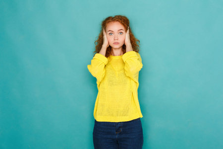 Young Woman Stands Against Blue Wall Covering Her Ears in a Yellow Sweater During Daytime
