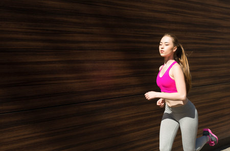 Woman Runs Outdoors in Fitness Attire During Daytime Near a Wooden Wall