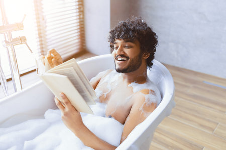 Man Reading a Book While Relaxing in a Bathtub Filled With Bubbles in a Bright Bathroom