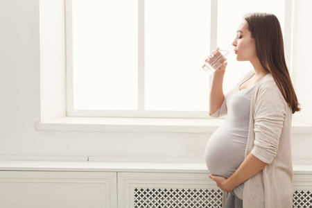 Pregnant Woman Drinking Water by a Window in a Bright Room With a Simple Design