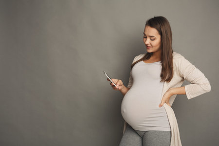 Pregnant Woman Checks Messages on Smartphone While Standing Against Gray Background