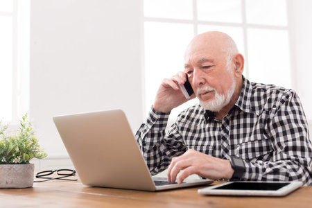 Elderly Man Speaks on the Phone While Working on a Laptop in a Bright Room During the Day