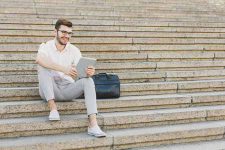 Man Sits on Steps and Uses Tablet While Smiling in a Public Area During Daytimeの写真素材
