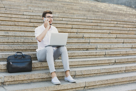 Man Sits on Steps Using Laptop While Talking on Phone in Outdoor Setting