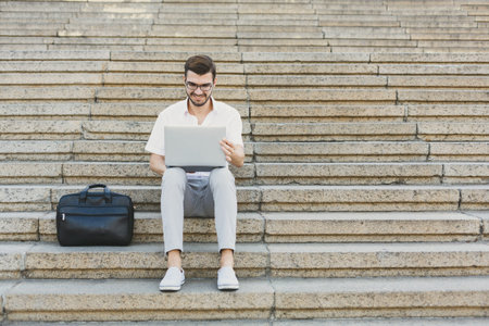 Young Man Sits on Steps With Laptop and Bag, Focused on Work or Study in an Outdoor Area