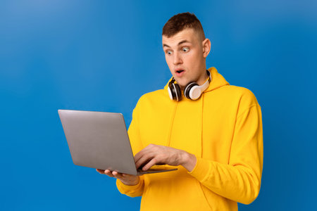 Emotional guy looking at laptop screen in shock, excited about win or success over blue background