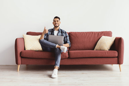 Man Sits on Couch With Laptop, Smiling and Giving Thumbs up in Modern Roomの写真素材