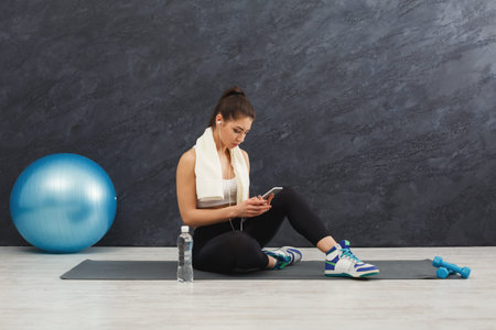 Woman Sitting on Mat Using Phone With Workout Gear and Equipment in a Gym or Fitness Studio