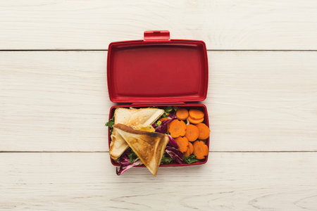 Lunch Set Displayed in a Red Lunchbox With Sandwich and Vegetables on Wooden Table
