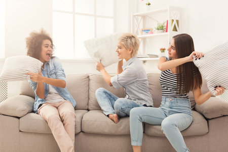 Friends Enjoy a Fun Pillow Fight in a Bright Living Room During a Weekend Gatheringの写真素材