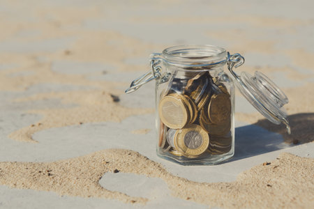 Collection of Coins in a Glass Jar on Sandy Surface Showing Texture and Light Reflections