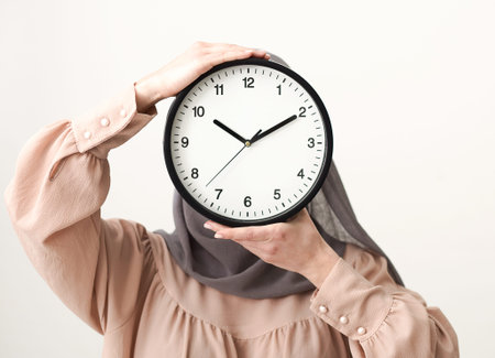 Woman Holds Clock Above Her Head in a Simple Setting While Wearing a Light-Colored Outfit and Hijab