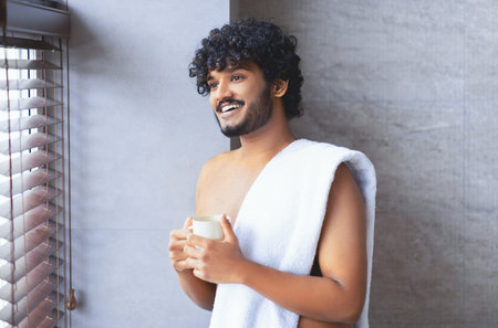Man Holding a Cup Standing Near a Window With Blinds in a Bathroom Setting