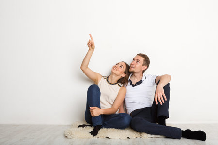 Couple Sitting on Floor and Looking up While Pointing at Blank Wall in Indoor Spaceの写真素材