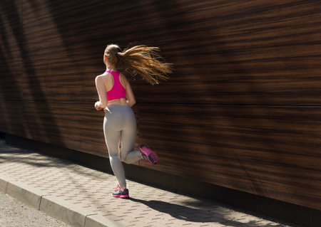 Woman Runs on the Sidewalk in Workout Clothes Near a Wooden Wall on a Sunny Day
