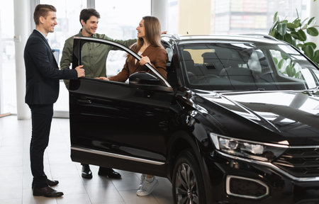 Cheery young couple speaking about buying new car with auto salesman at vehicle dealership