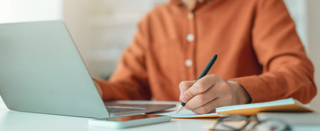 Person Works on Laptop While Taking Notes at a Desk in a Bright Room During the Day