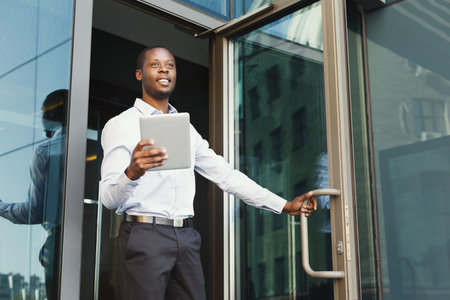 Man Steps Through a Glass Door While Holding a Tablet in His Hand During a Sunny Day