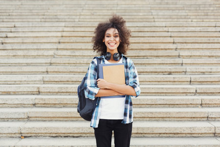 Smiling Young Student Stands on Steps Holding Books and Wearing Headphones in a Casual Setting