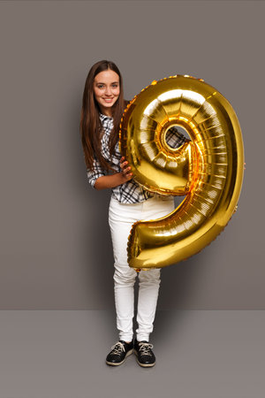 A Girl Stands in a Studio Setting, Holding a Large Gold Balloon Shaped Like the Number Nine