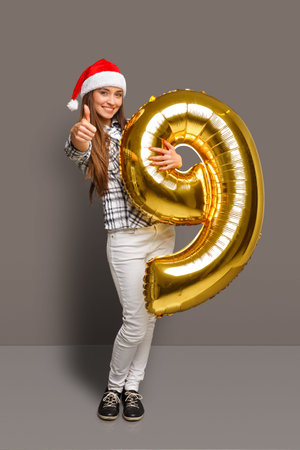 Young Person Celebrates Holiday With Golden Number Balloon While Wearing a Santa Hat