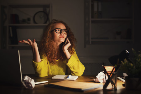 Woman Talking on Phone in Dark Room With Clutter on Desk and Look of Frustration
