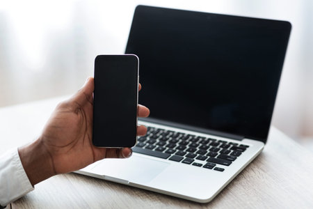Black man holding smartphone with blank screen, while using laptop