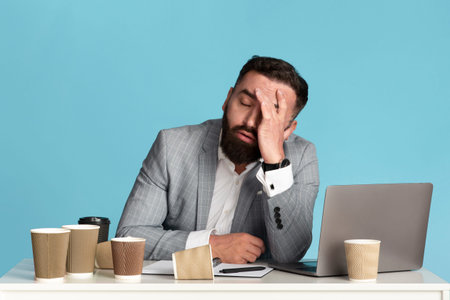 Working overtime. Exhausted businessman sitting at desk with laptop and empty coffee cups against blue background
