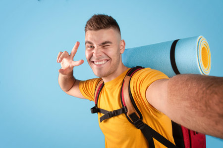Funky millennial hipster with camping equipment taking selfie, winking and showing peace gesture over blue background