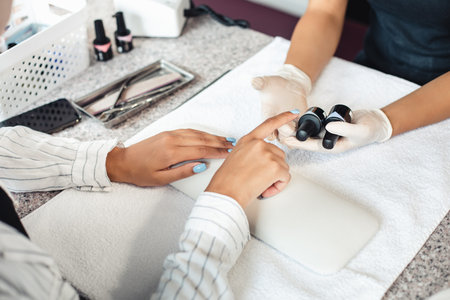 Manicure and pedicure salon with security measures in reopening of pandemic. African american woman client chooses color of varnish in hands of master in rubber gloves on table