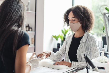 Manicure procedure in studio and social distance during covid-19 epidemic. Woman in rubber gloves covers nails with blue polish to african american lady in protective mask