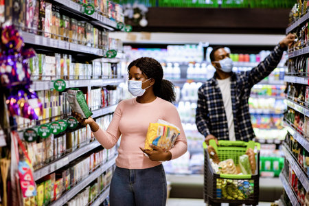Young black woman and man in face masks choosing food products at supermarket during covid quarantine