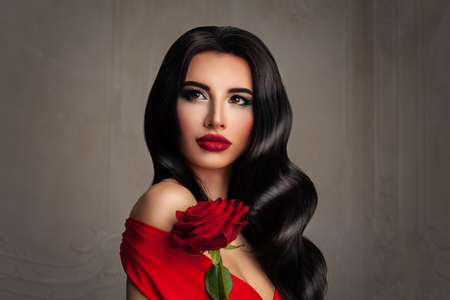 Beautiful Lady. Fashion Portrait of Perfect Woman with Brunette Hair and Event Makeup. Curly Hairstyle, Red Lips, and Red Rose Flowerの写真素材
