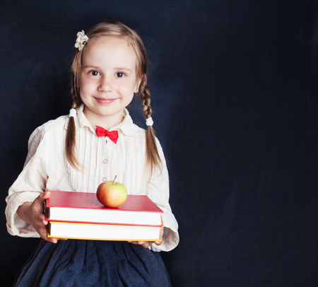 Cute little schoolgirl with red apple and books. Happy child on empty blackboard background with copy space. Back to school.の写真素材