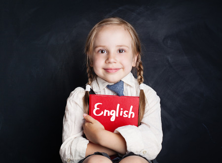English language concept. Cute child pupil with book smiling and sitting in classroom against blackboard backgroundの写真素材