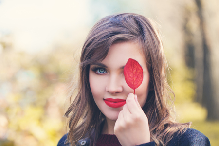 Autumn woman with red lips makeup holding red fall leaf in autumn park, portraitの写真素材