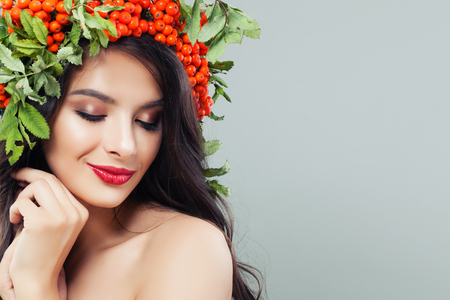 Beautiful female face closeup. Brunette woman with long wavy hair, makeup and red berries and green leaves wreath on banner background with copy spaceの写真素材
