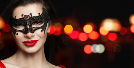 Perfect woman face in black carnival mask and red lips makeup on black background with abstract night glitter bokehの写真素材