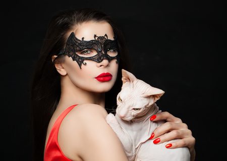 Sexy brunette woman with red lips makeup, carnival mask and hairless cat on black background, Halloween conceptの写真素材
