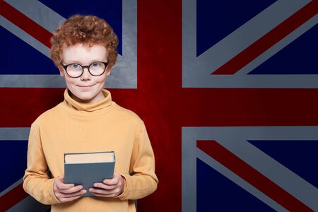Smart kid holding book against the UK flag background. Learn English conceptの写真素材