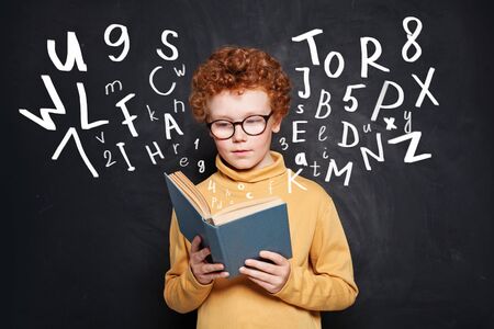 Curious little boy in glasses reading a book against chalkboardの写真素材