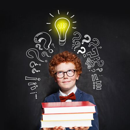 Little boy wearing bow tie and suit with lightbulb and books on blackboard background. Brainstorming and idea conceptの写真素材