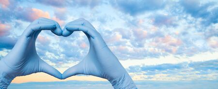 Doctor hands in medica gloves showing heart on sky clouds backgroundの写真素材