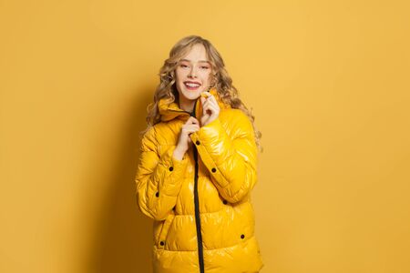 Young happy cheerful fashion model woman in winter jacket smiling on bright yellow background