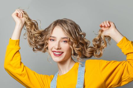 Young cheerful blonde woman touching her long curly hair on gray backgroundの写真素材