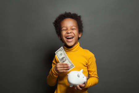 Smart happy black child boy putting in piggy bank one dollars banknoteの写真素材