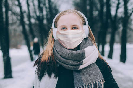 Young girl in medical protective face mask walking in winter park and listening music with wireless headphonesの写真素材