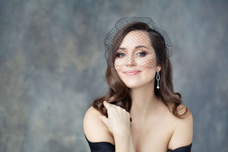Elegant brunette model woman with long curly hair in black veil on gray stucco background